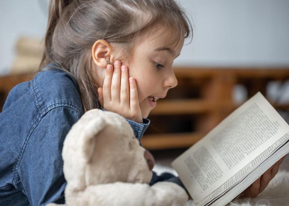 Little girl reads a book with a teddy bear.